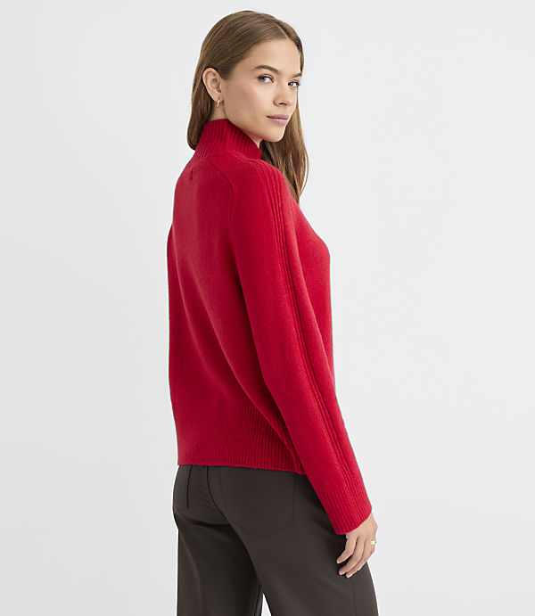 A woman in a red turtleneck sweater and dark pants stands against a plain gray background.