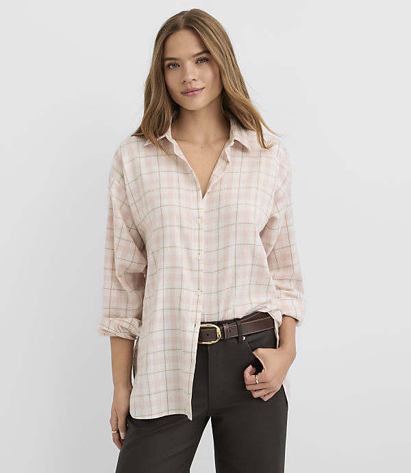A woman in a light pink plaid shirt and dark pants stands against a plain white background.