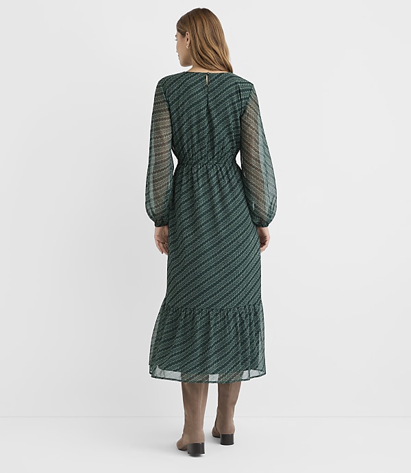 A woman in a green patterned dress and brown boots stands against a plain white background.