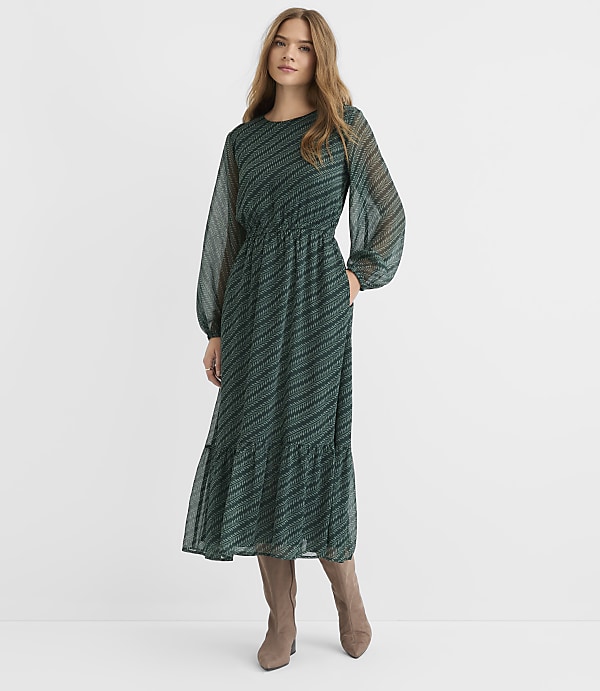 A woman in a long, green, patterned dress stands against a plain, light gray background.