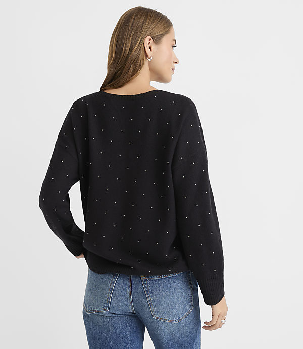 A woman in a black studded sweater and blue jeans stands against a plain white background.