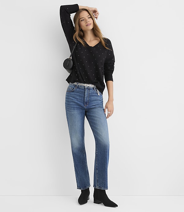 A woman in a black embellished sweater and blue jeans stands against a plain white background.