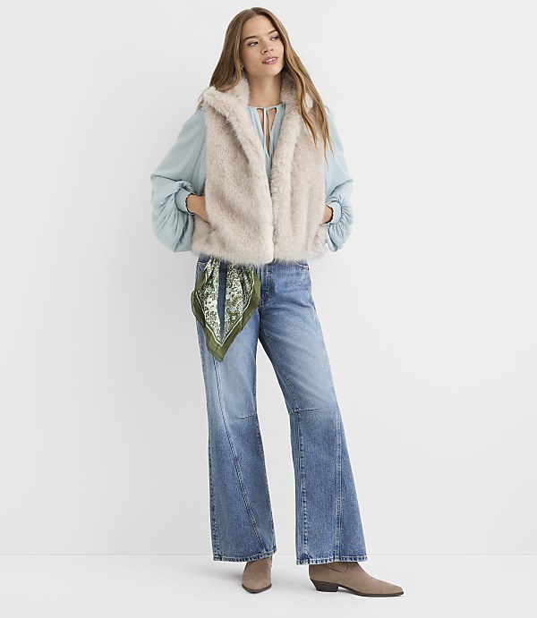 A woman in a faux fur vest and wide-leg jeans stands against a plain white background.