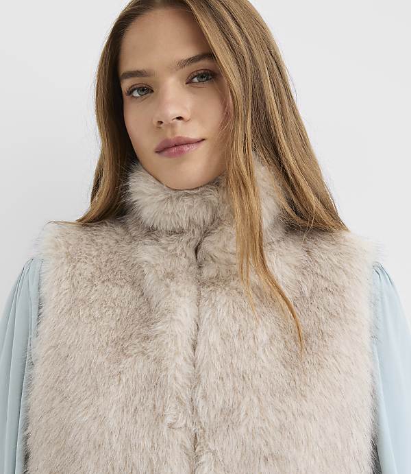 A woman with long blonde hair wears a light beige fur vest over a light blue blouse against a plain white background.