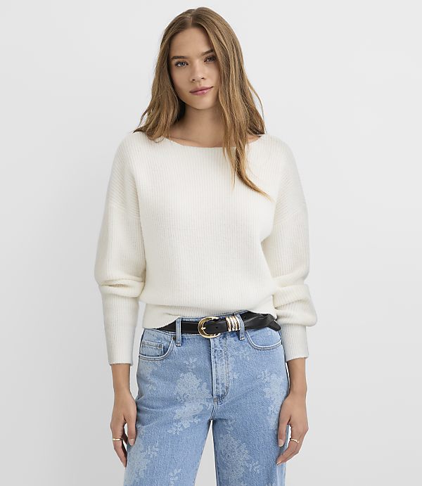 A woman in a white sweater and patterned jeans stands against a plain white background.