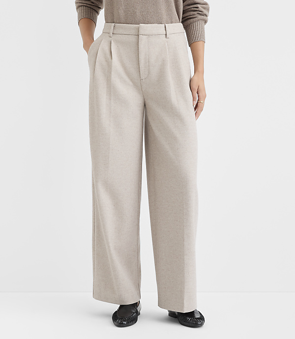 A person stands in the foreground wearing beige pleated wide-leg pants and a brown sweater, against a plain white background.