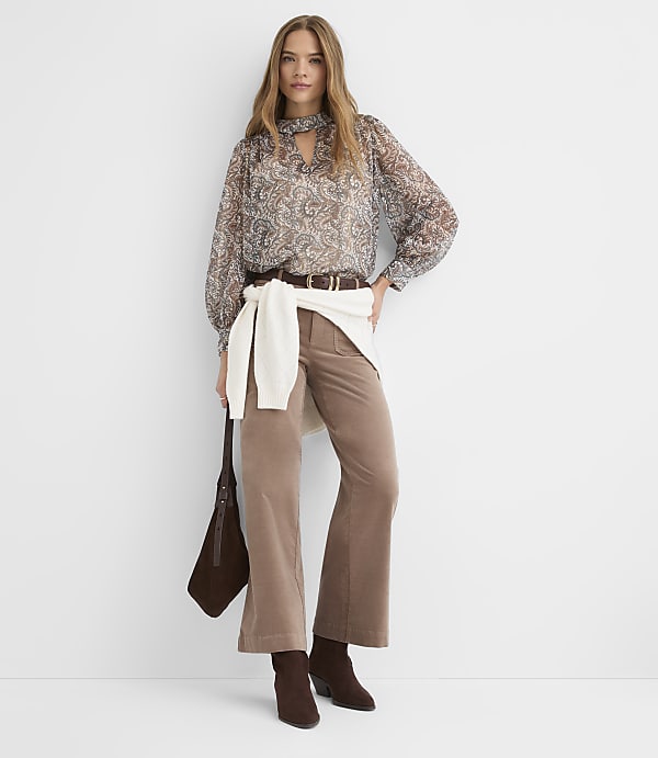 A woman in a patterned blouse and brown corduroy pants stands against a plain white background, holding a brown purse.