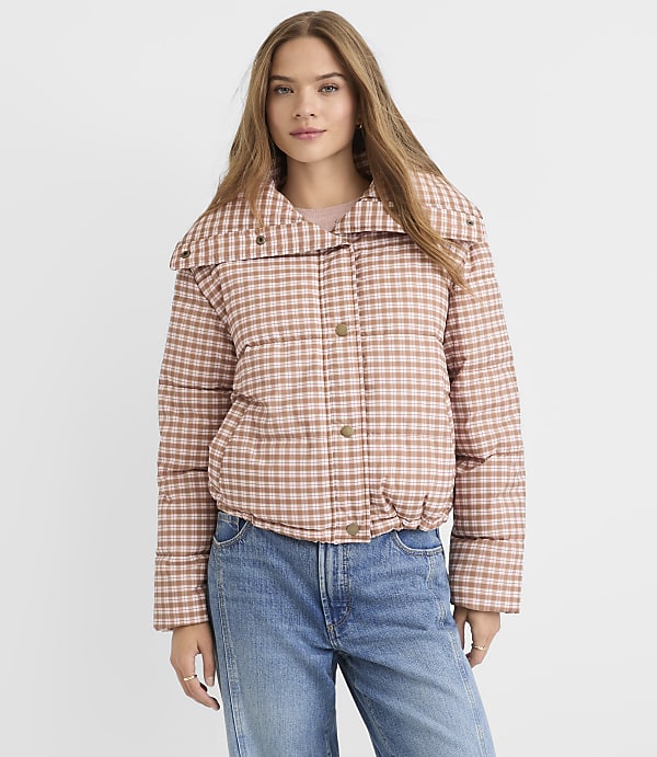 A young woman in a pink and brown checkered puffer jacket and blue jeans stands against a plain white background.