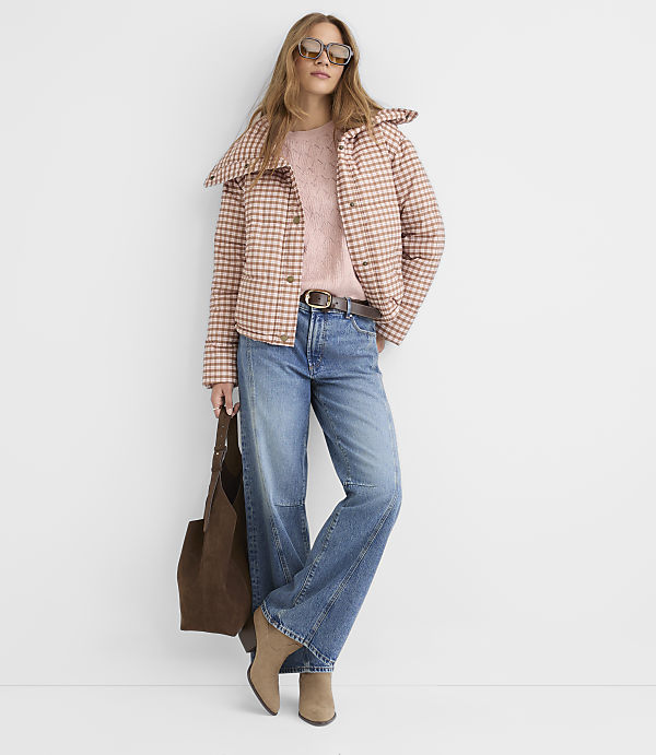 A woman in a pink plaid jacket and jeans stands against a plain white background, holding a brown tote bag.