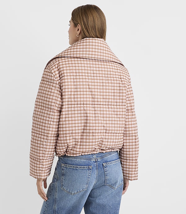 A woman in a pink gingham puffer jacket and blue jeans stands against a plain white background.