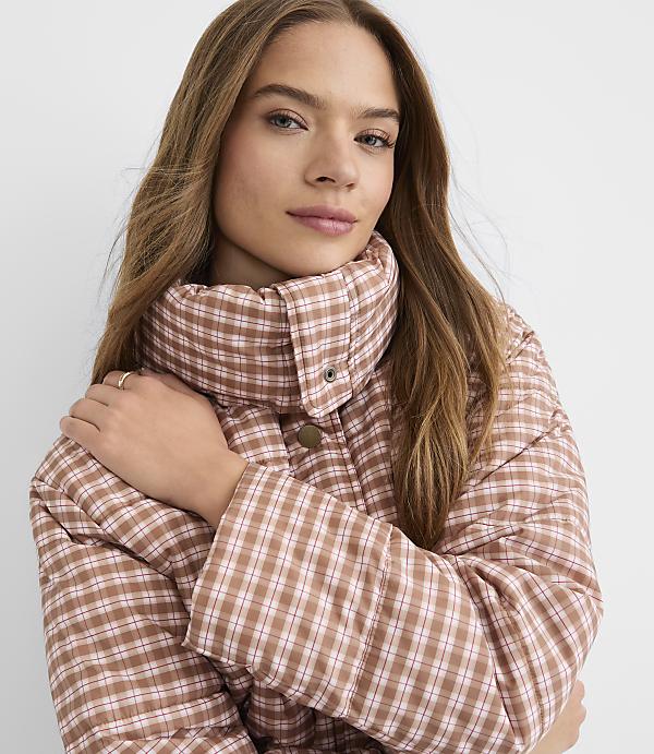 A young woman with long, wavy brown hair wears a brown and white plaid puffer jacket with a high collar, set against a plain white background.