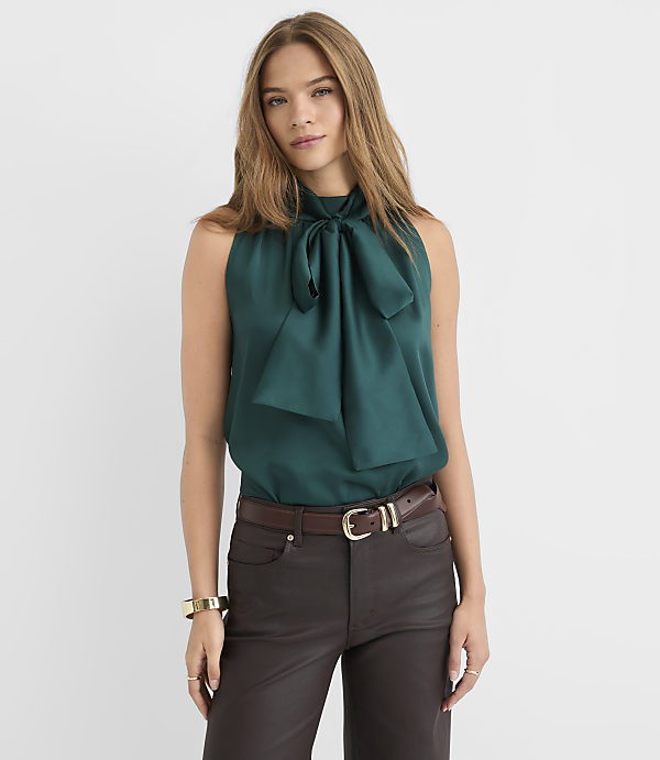 A woman in a teal bow-neck blouse and dark pants stands against a plain white background.