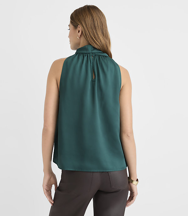 A woman in a teal sleeveless top and brown pants stands against a plain white background.