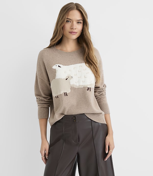 A woman in a tan sweater with sheep appliqu&eacute;s stands against a plain, light background.
