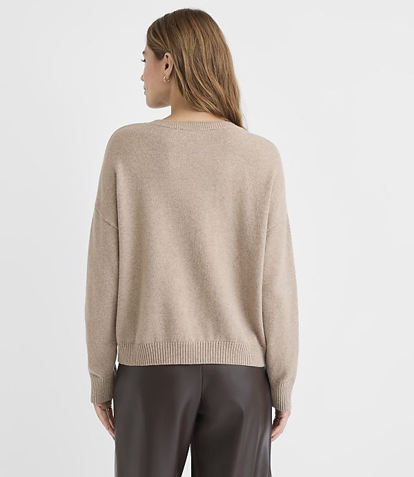 A woman in a beige sweater and brown leather pants stands against a plain white background.