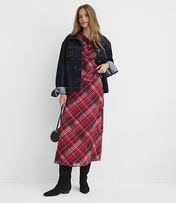 A woman in a plaid dress and denim jacket stands against a plain white background.