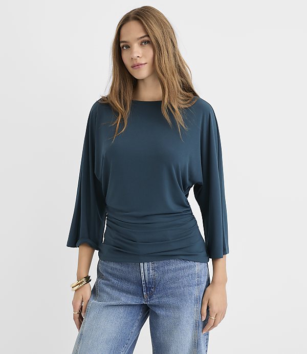 A woman in a teal top and jeans stands against a plain white background.