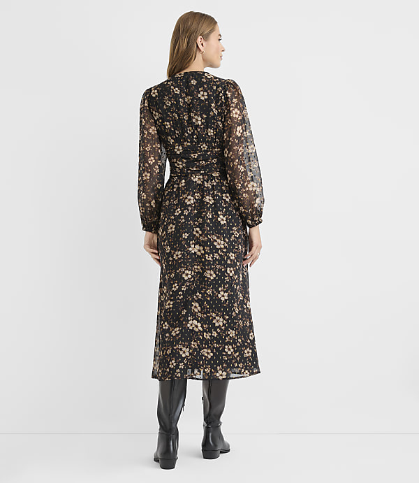 A woman in a floral midi dress and black boots stands against a plain, light gray background.