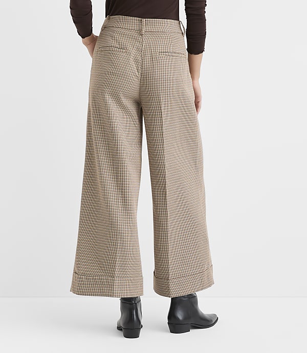 A person stands in the foreground wearing wide-leg, checkered pants and black boots, with a plain brown background.
