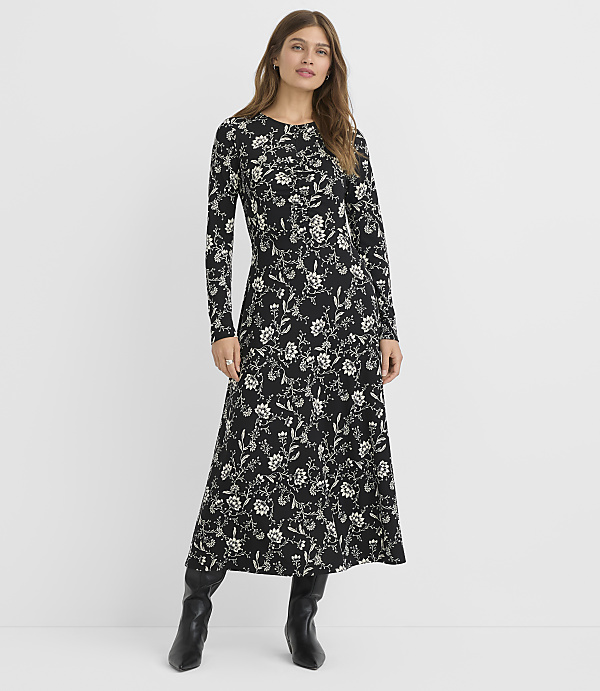 A woman in a black floral dress and black boots stands against a plain white background.