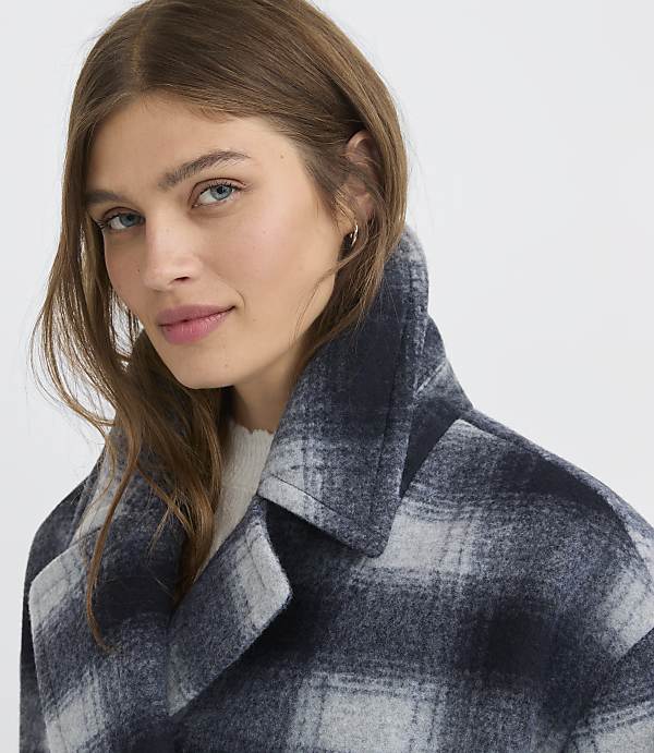 A woman with brown hair and blue eyes wears a plaid coat in the foreground, against a plain white background.