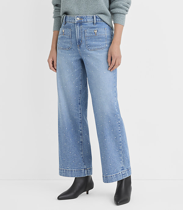 A person stands in light-wash, wide-leg jeans adorned with small white embellishments, against a plain white background.