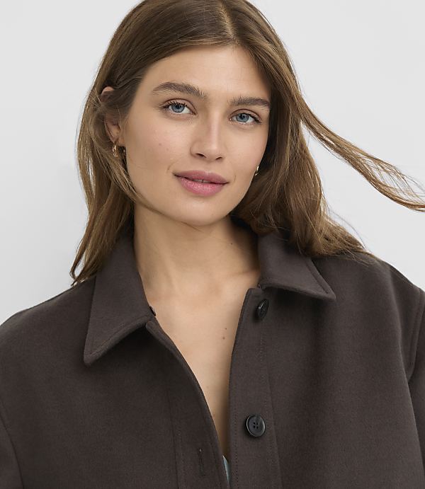 A young woman with long brown hair and light blue eyes smiles gently, wearing a dark brown collared jacket against a plain white background.