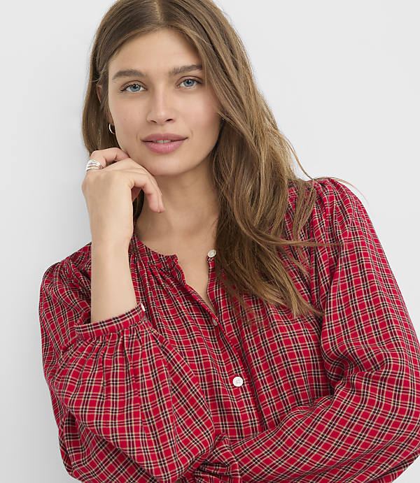 A woman with long brown hair and a red plaid shirt rests her chin on her hand against a plain white background.