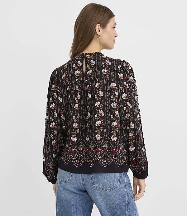 A woman in a patterned blouse and jeans stands against a plain gray background.