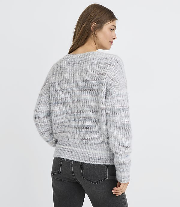 A woman in a grey speckled sweater and dark jeans stands against a plain white background.