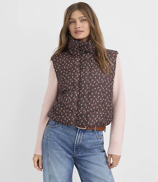 A woman in a floral puffer vest and pink sweater stands against a plain background.