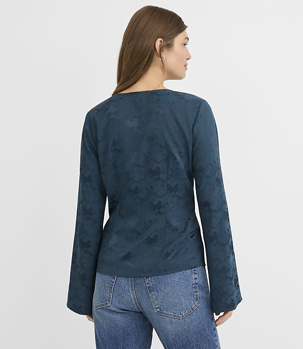 A woman in a teal patterned long-sleeved top and blue jeans stands against a plain white background.