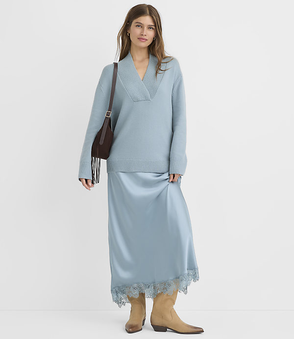 A woman in a light blue sweater and satin skirt with lace trim stands against a plain white background, carrying a brown fringed crossbody bag and wearing tan cowboy boots.