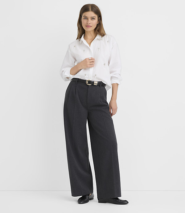 A woman in a white button-down shirt and wide-leg gray pants stands against a plain white background.
