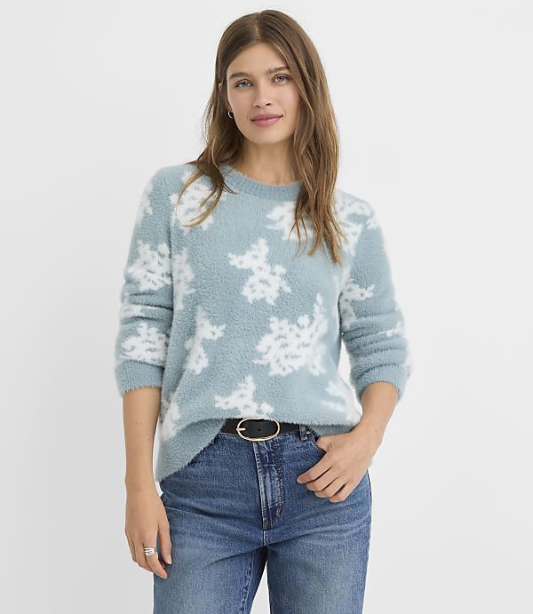 A woman in a fuzzy blue sweater with a floral pattern and jeans stands against a plain, light background.