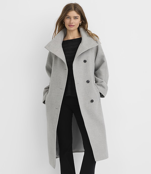A woman in a gray coat stands against a plain white background.