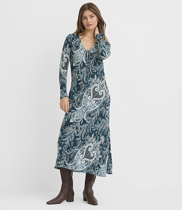 A woman in a long, patterned dress and brown boots stands against a plain white background.