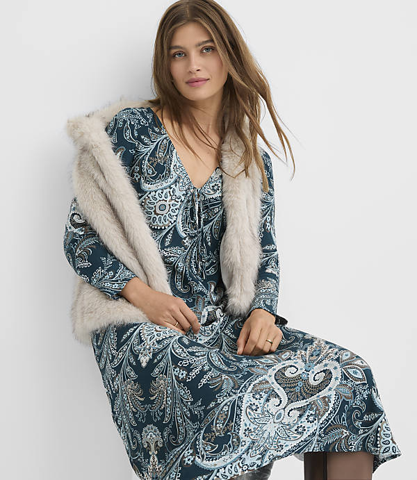 A woman in a teal paisley dress and a faux fur stole poses against a plain white background.