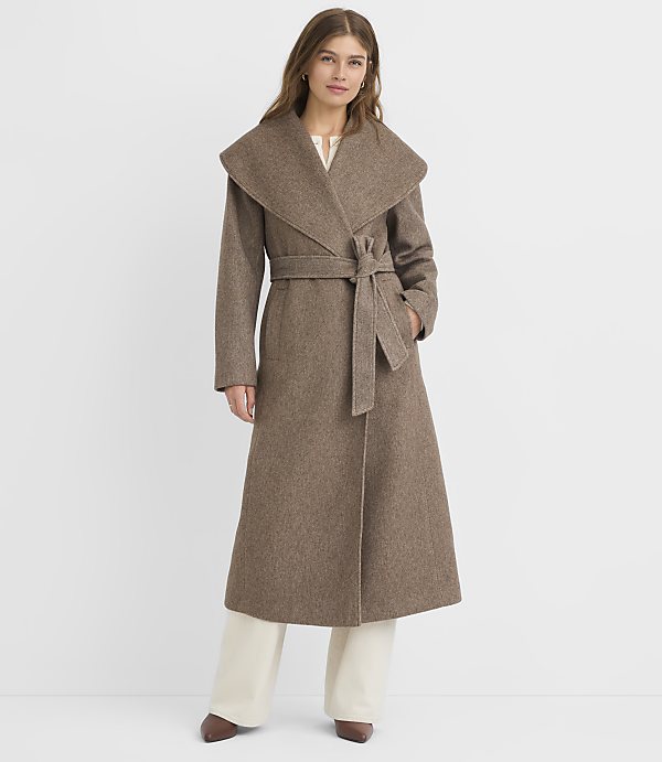 A woman in a long, brown, belted coat stands against a plain white background.