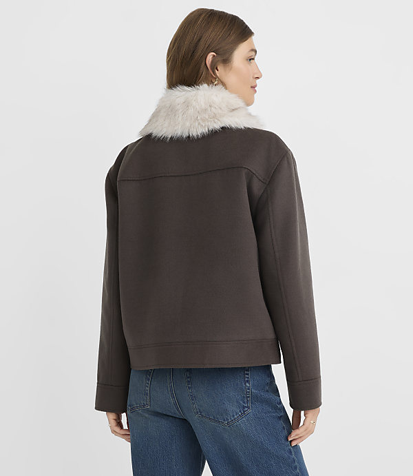 A woman in a brown jacket with a fur collar and blue jeans stands against a plain white background.
