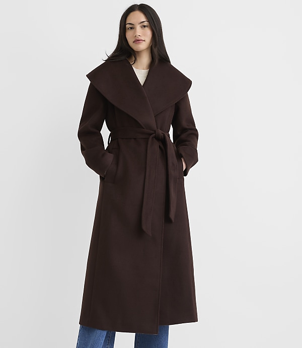 A woman in a long brown coat stands against a plain white background.