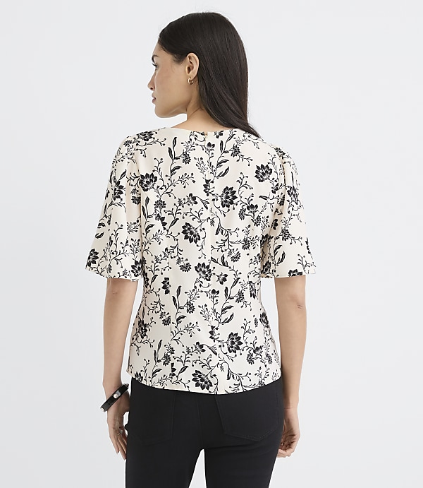 A woman in a cream-colored floral blouse and black pants stands against a plain white background.