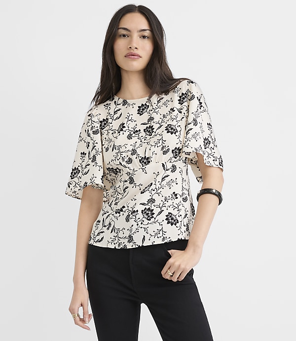 A woman in a floral top and black pants stands against a plain white background.