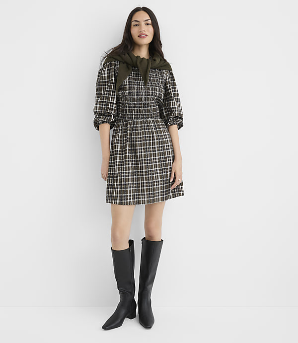 A woman in a plaid dress and black boots stands against a plain white background.