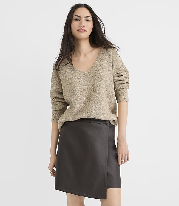 A woman in a tan sweater and dark skirt stands against a plain gray background.