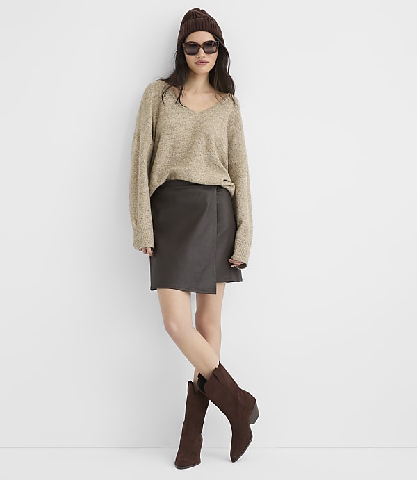 A woman in a beige sweater, brown beanie, sunglasses, and a dark skirt stands against a plain white background, with her legs crossed and wearing brown boots.