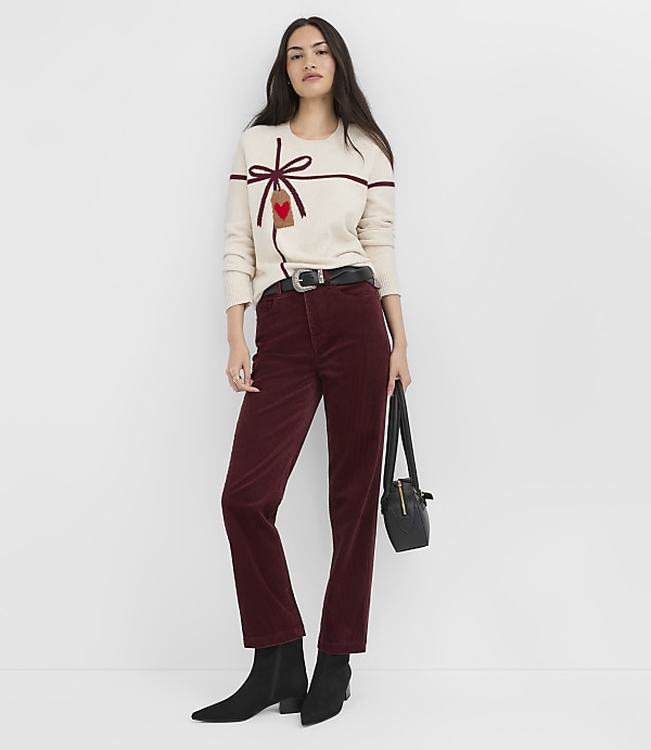 A woman in a cream sweater and burgundy pants stands against a plain white background, holding a black purse.