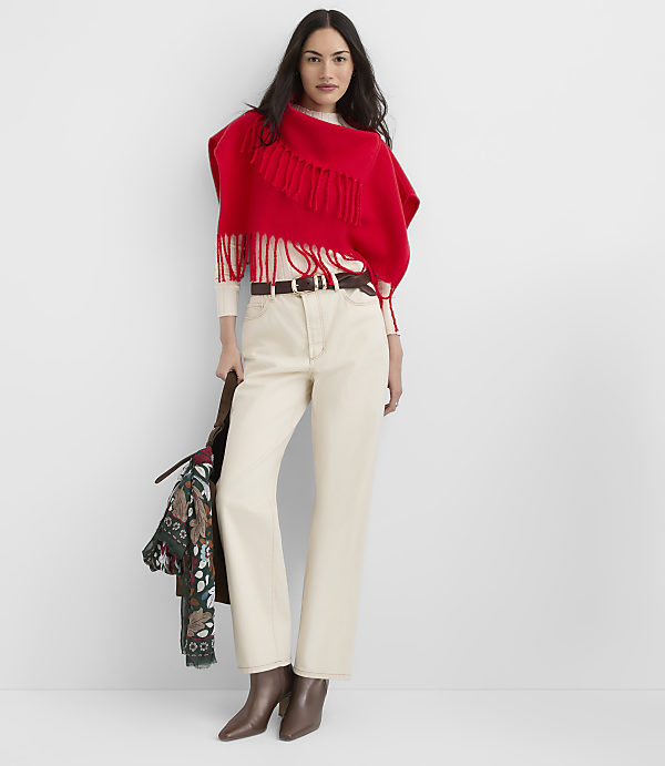A woman in a red fringed poncho and cream pants stands against a plain white background, holding a patterned bag.