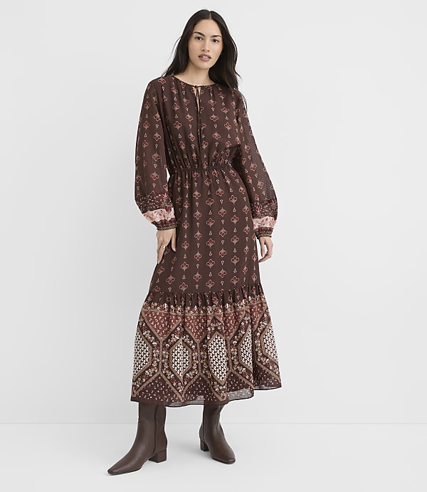 A woman in a long, patterned brown dress and boots stands against a plain white background.