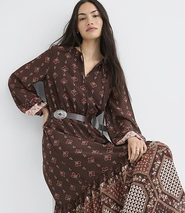 A woman in a patterned brown dress with a belt stands against a plain white background.